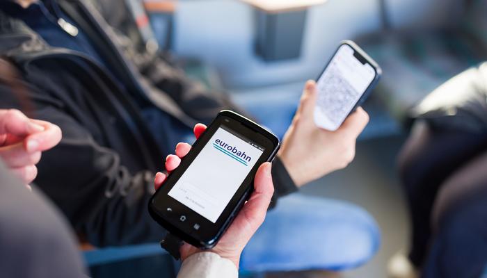 Eine Fahrkartenkontrolle, wobei die Detailaufnahme die Hand des Kundenbetreuers, die ein Lesegerät hält, zeigt. Auf dem Display des Handys des Gastes ist die digitale Fahrkarte zu sehen.